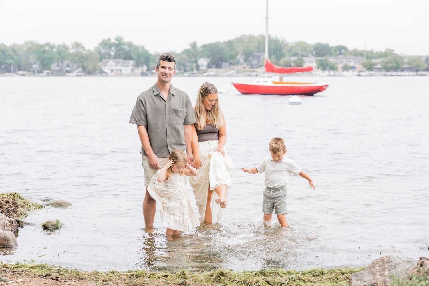 Family of four wading in shallow water near a rocky shore with a red sailboat in the background.