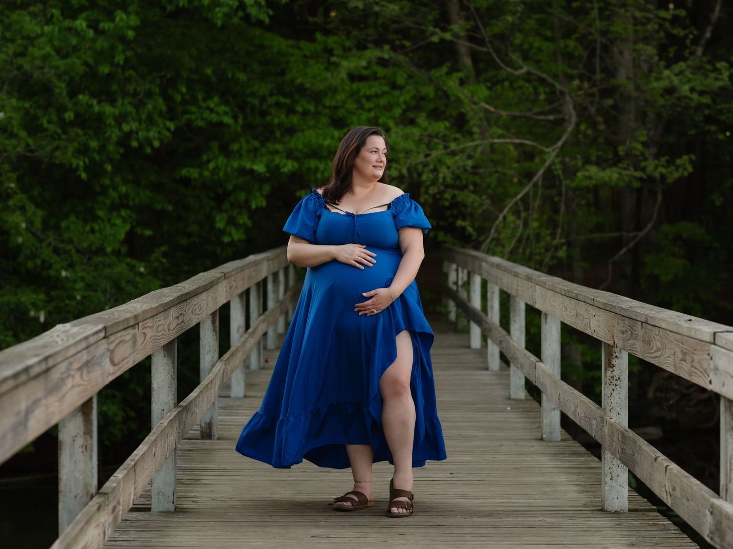 Pregnant woman in blue off-shoulder dress posing on wooden bridge surrounded by lush green trees.