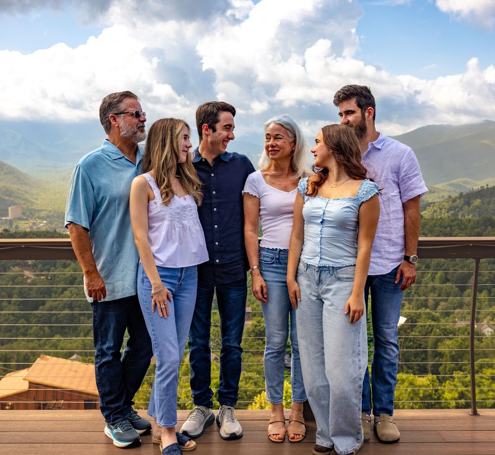 Family of six smiling together on a deck with scenic mountain views and cloudy sky backdrop.