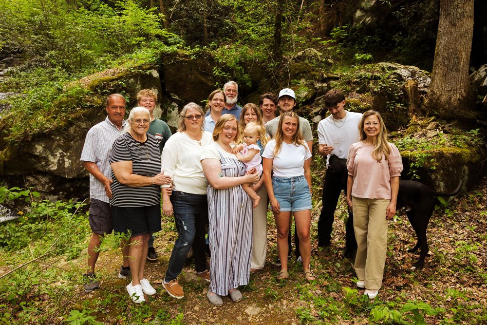 Large family group smiling together outdoors in a lush green forest setting near rocky terrain.