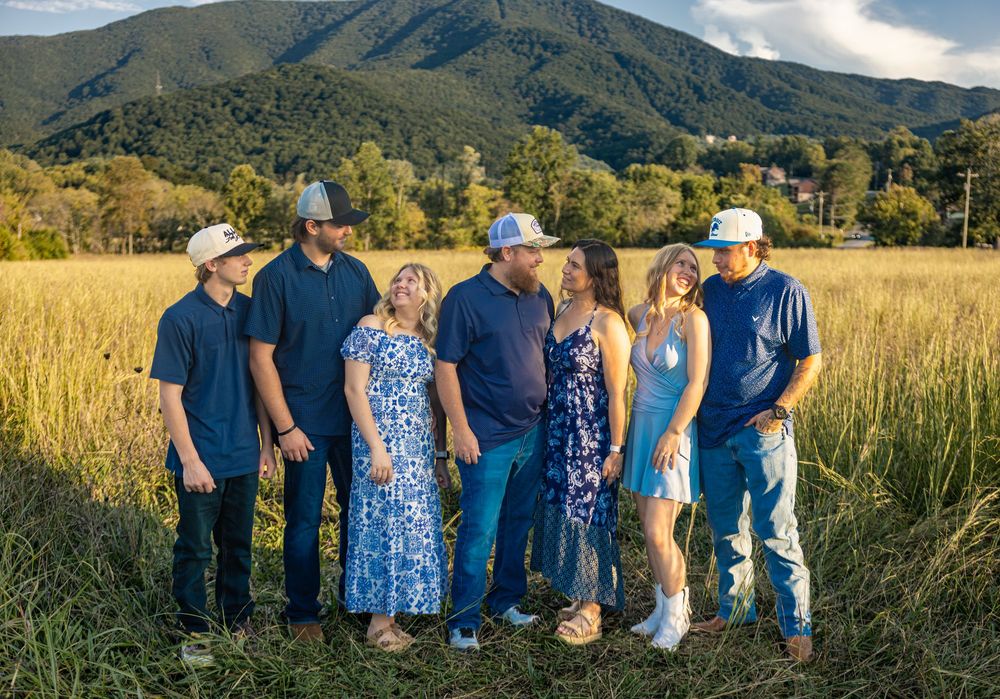 Family of seven posing in golden field with mountain backdrop, wearing casual country attire including cowboy hats.