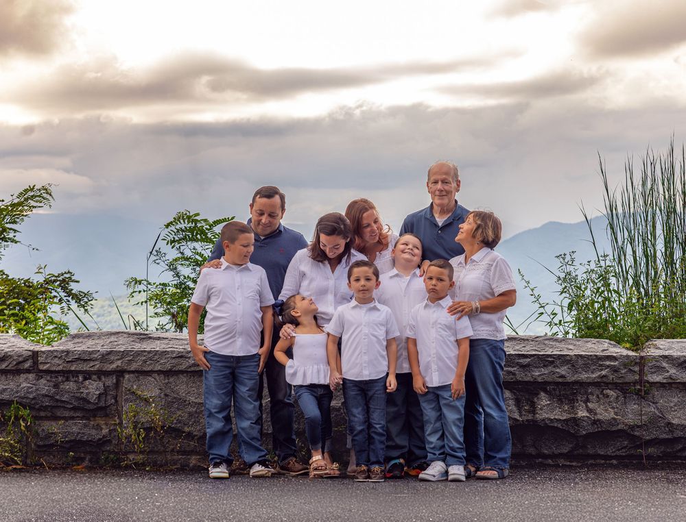 Large family group portrait outdoors with mountains and dramatic sky, adults and children in white shirts and jeans.