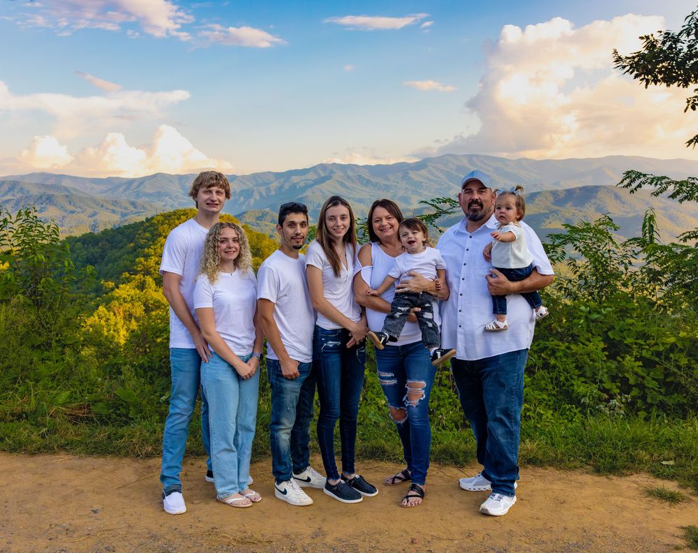 Family of seven posing together on a scenic mountain overlook with lush green hills and cloudy sky backdrop.
