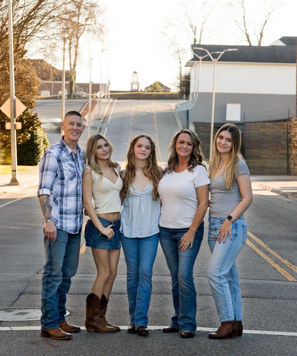 Family of five posing together on a small town street, smiling in casual outfits including jeans and plaid.