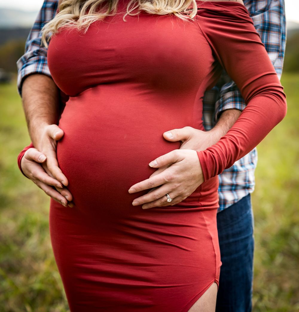 Pregnant woman in red dress with partner's hands on baby bump during outdoor maternity photo session.