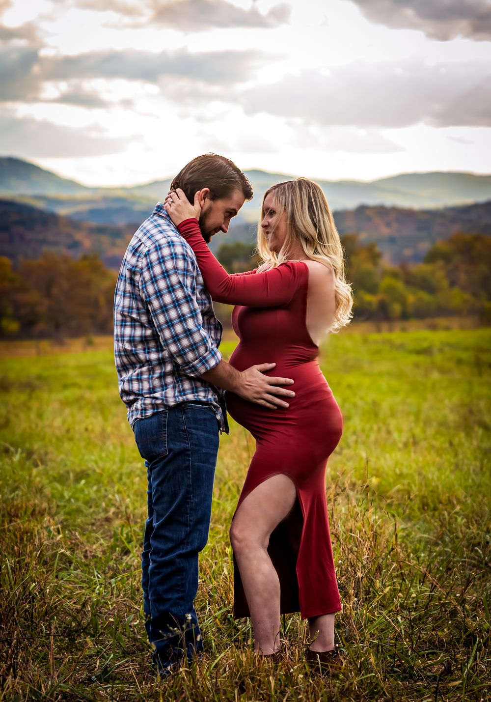 Couple embracing in a green field with rolling mountains backdrop, woman in red dress, man in plaid shirt and jeans.