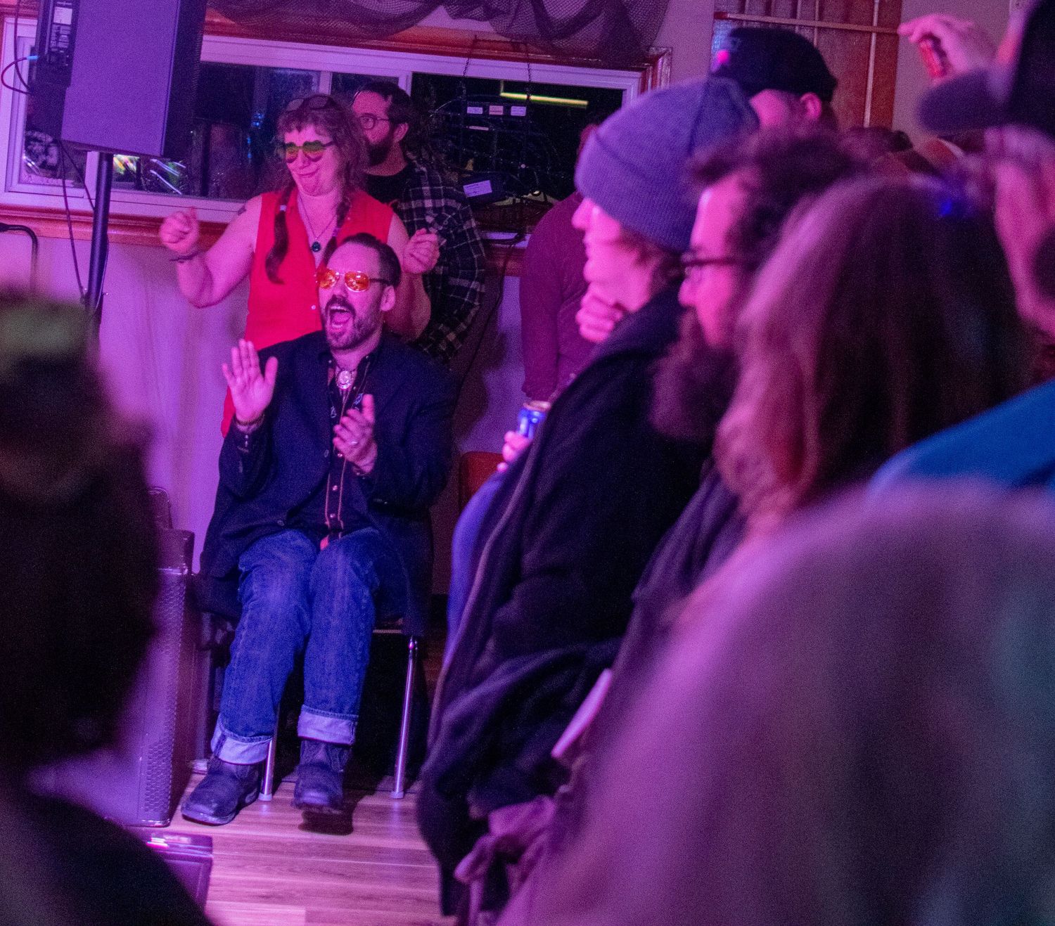 Animated speaker seated in chair addresses engaged crowd at intimate indoor event venue.