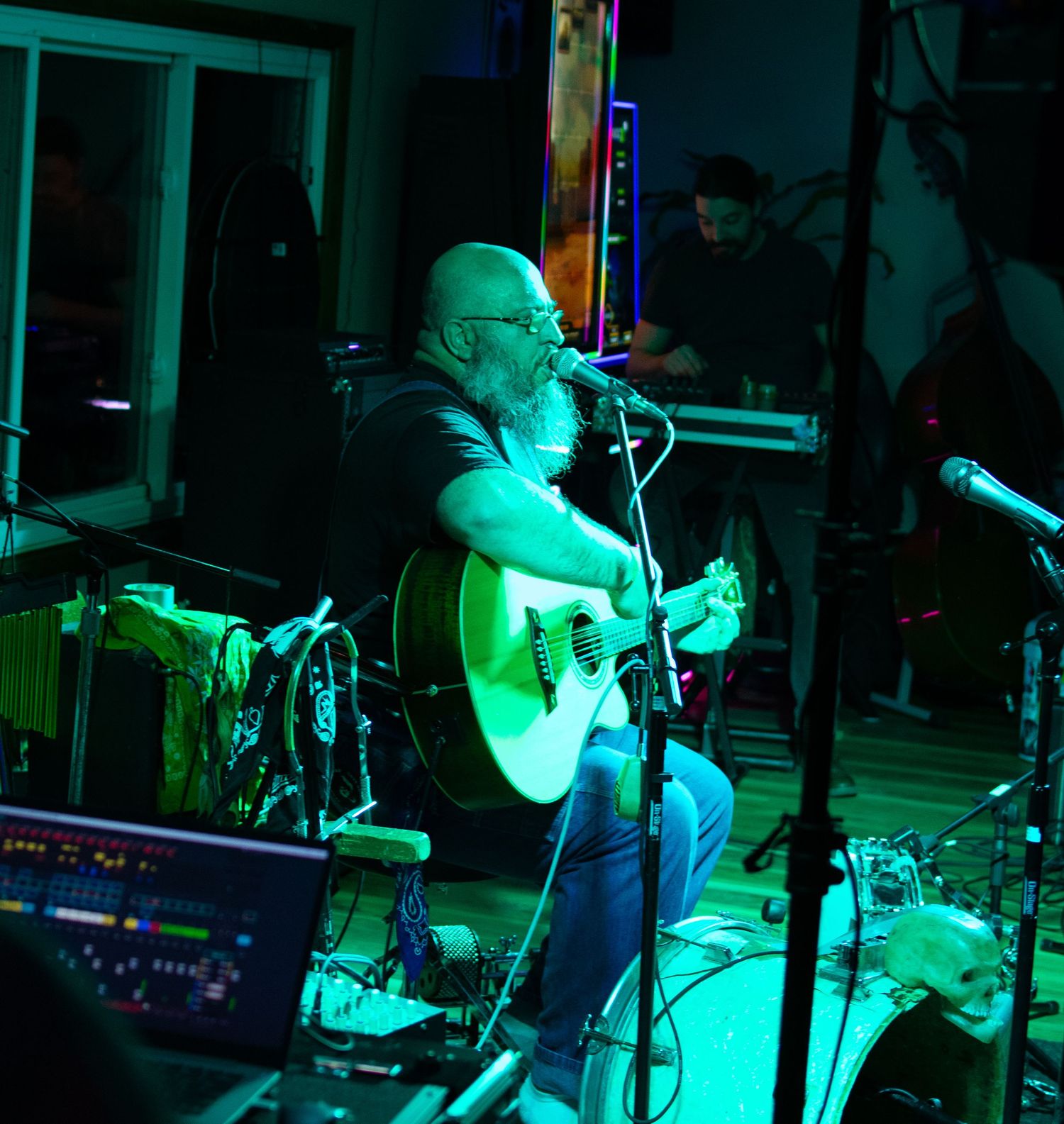Bald bearded guitarist performing acoustic set on stage under colorful venue lighting.
