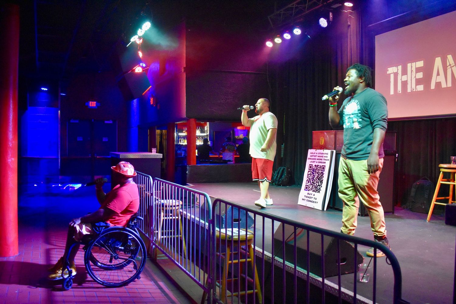 Three performers on colorful stage, one seated in wheelchair, at live entertainment venue.