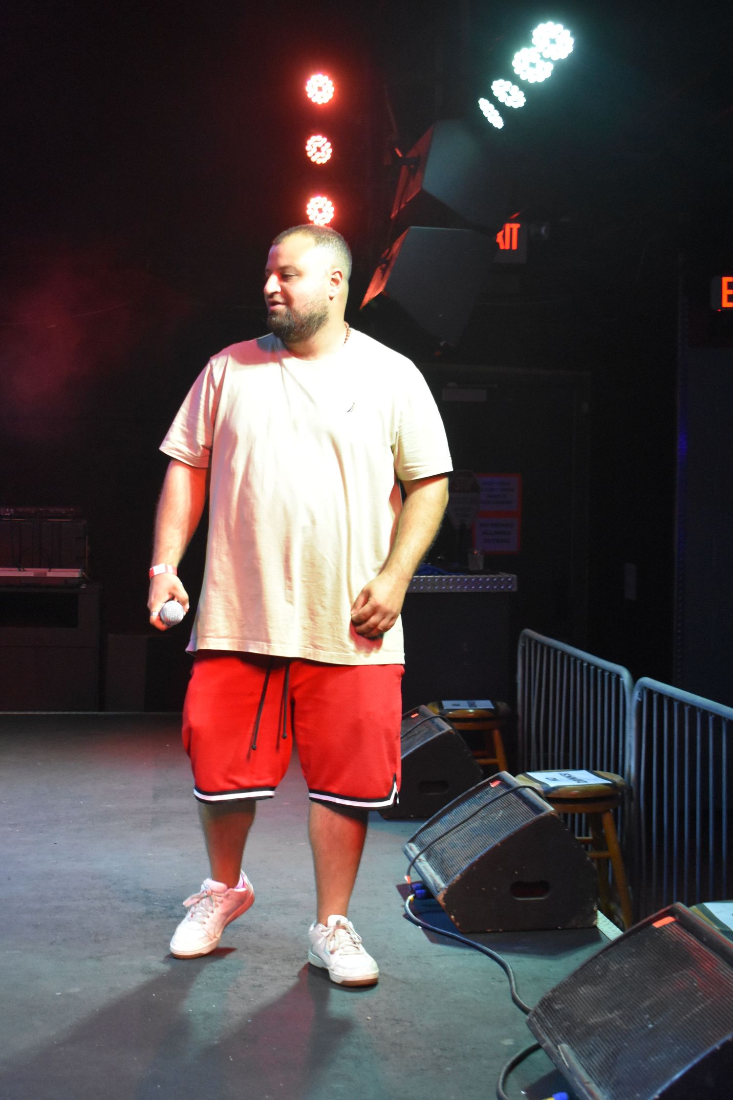 A heavyset man performs on stage wearing a white t-shirt and red shorts under bright stage lights.
