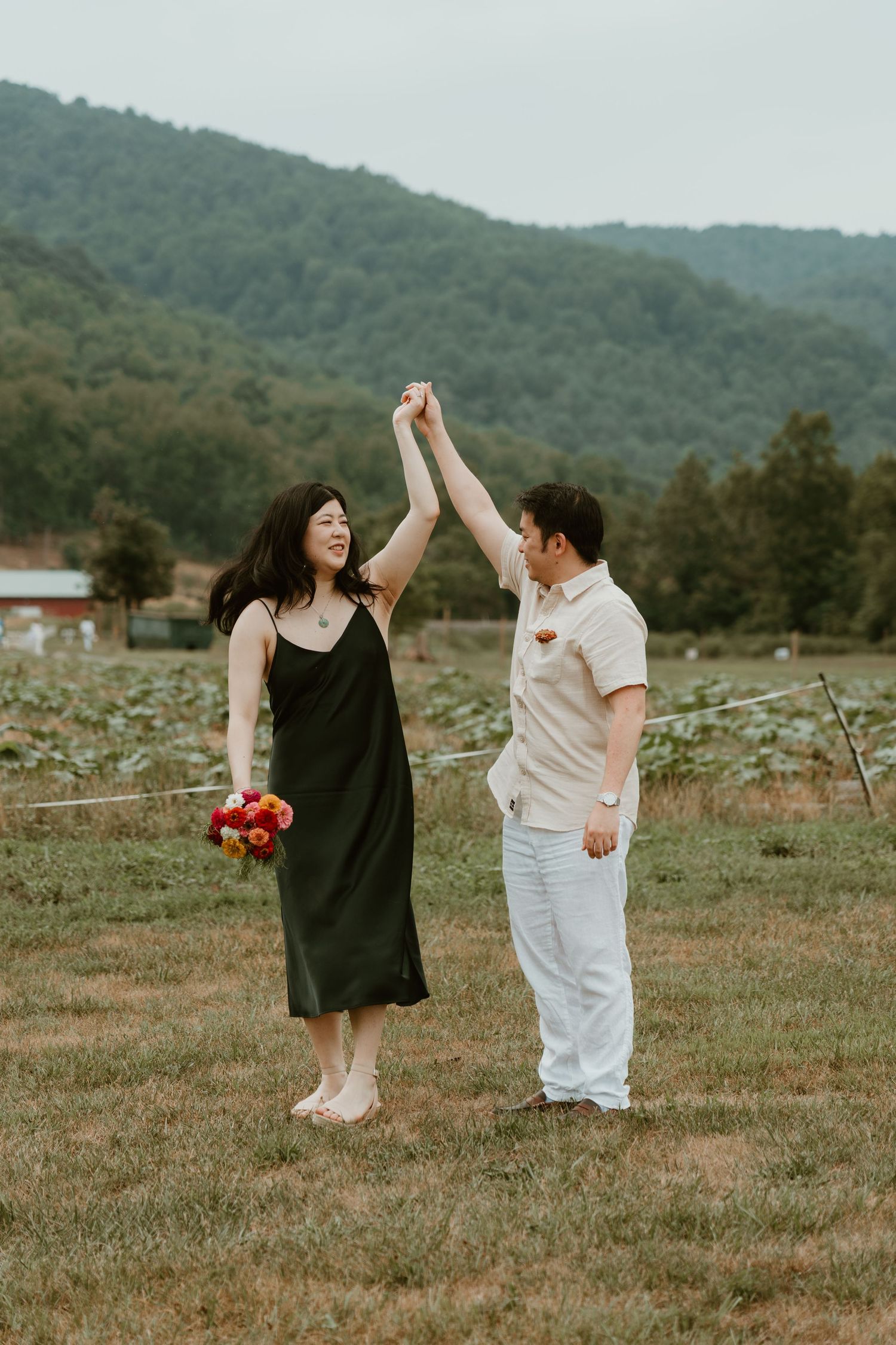 A couple dances joyfully in a green farm field with mountain backdrop, woman in black dress holding colorful bouquet.