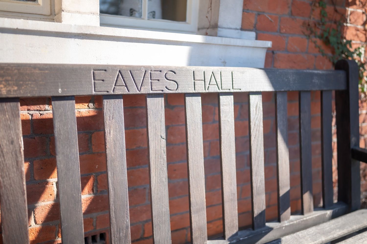 Wooden bench with carved Eaves Hall sign against a red brick building wall in natural sunlight.