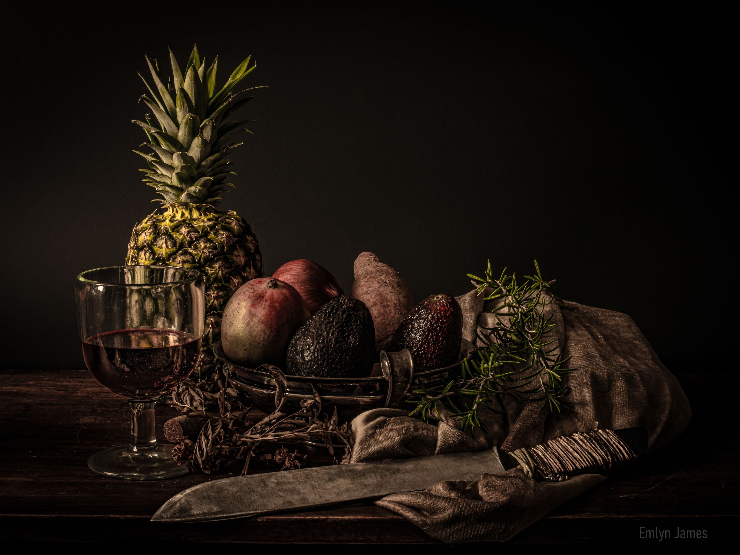 Dark moody still life with pineapple, avocados, peaches, rosemary, and a wine glass on a rustic wooden table.