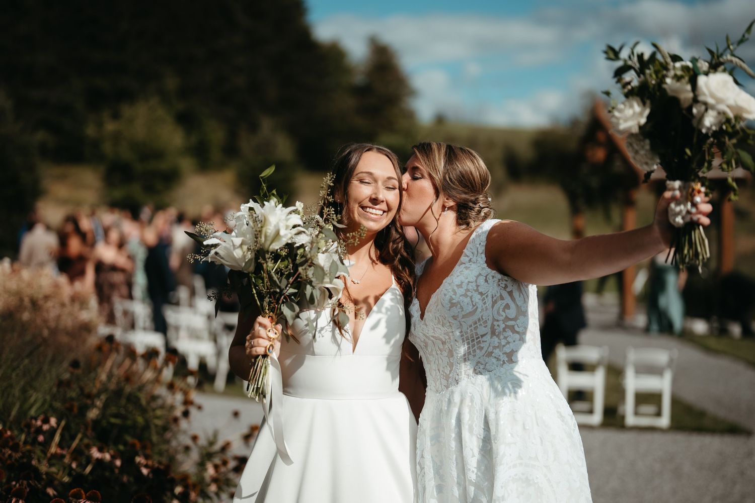Two brides in white wedding gowns share a kiss while holding bouquets outdoors at a garden wedding ceremony.