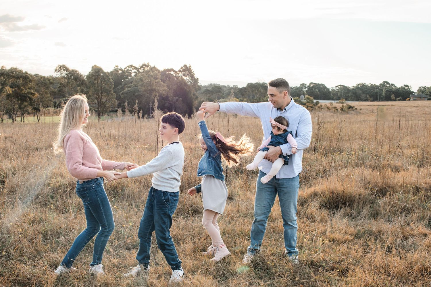 Family of four playing and dancing together in a golden field in Rouse Hill Regional Park at sunset, parents twirling children around.