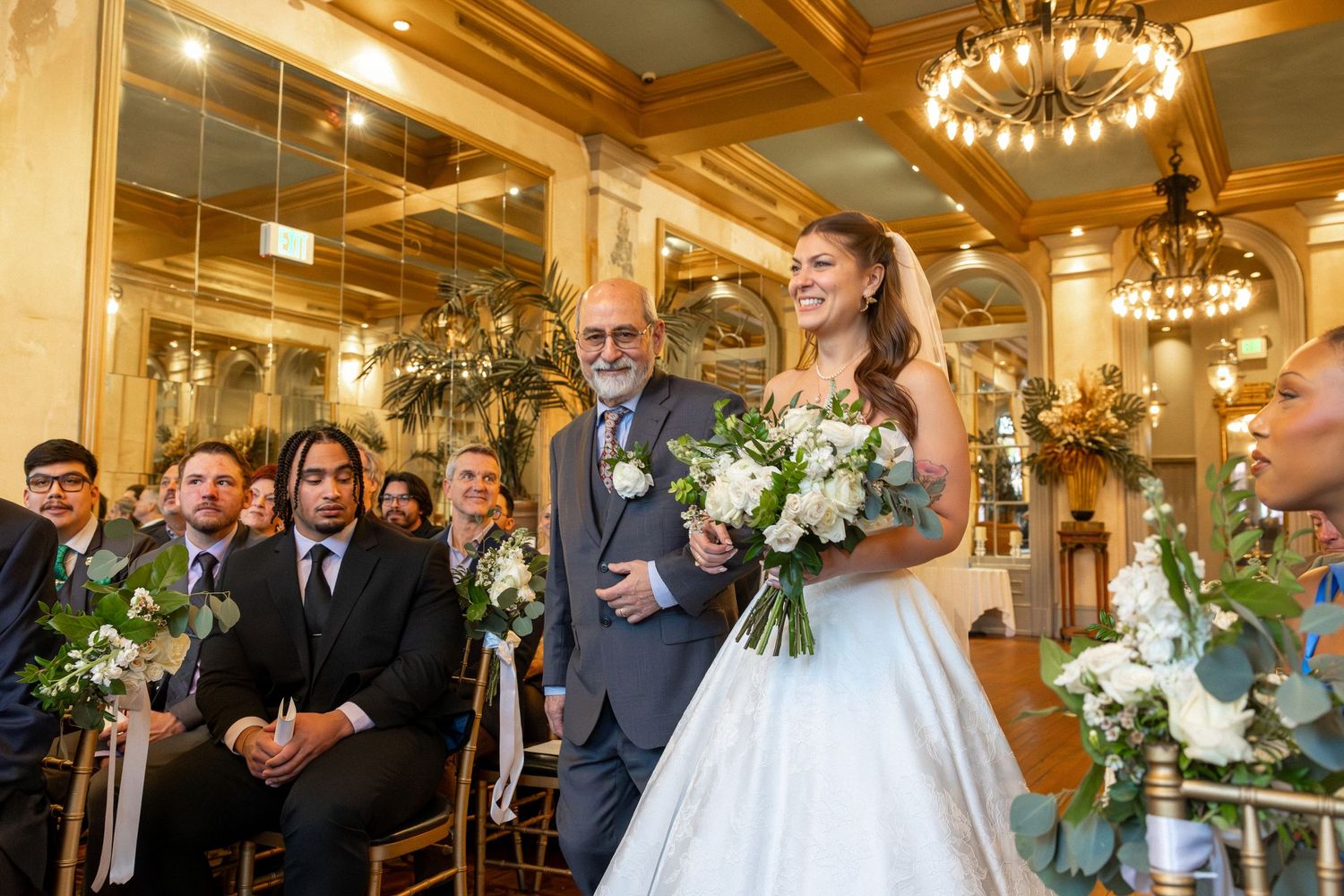 Bride in white gown walks down the aisle with her father at an elegant indoor wedding ceremony.