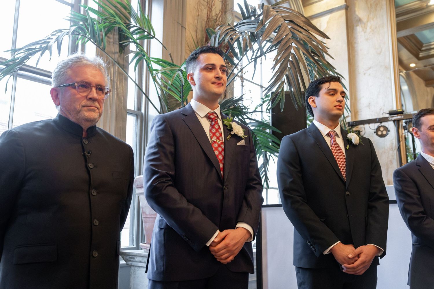 Three men in formal suits standing together at a wedding ceremony indoors near a large tropical plant.