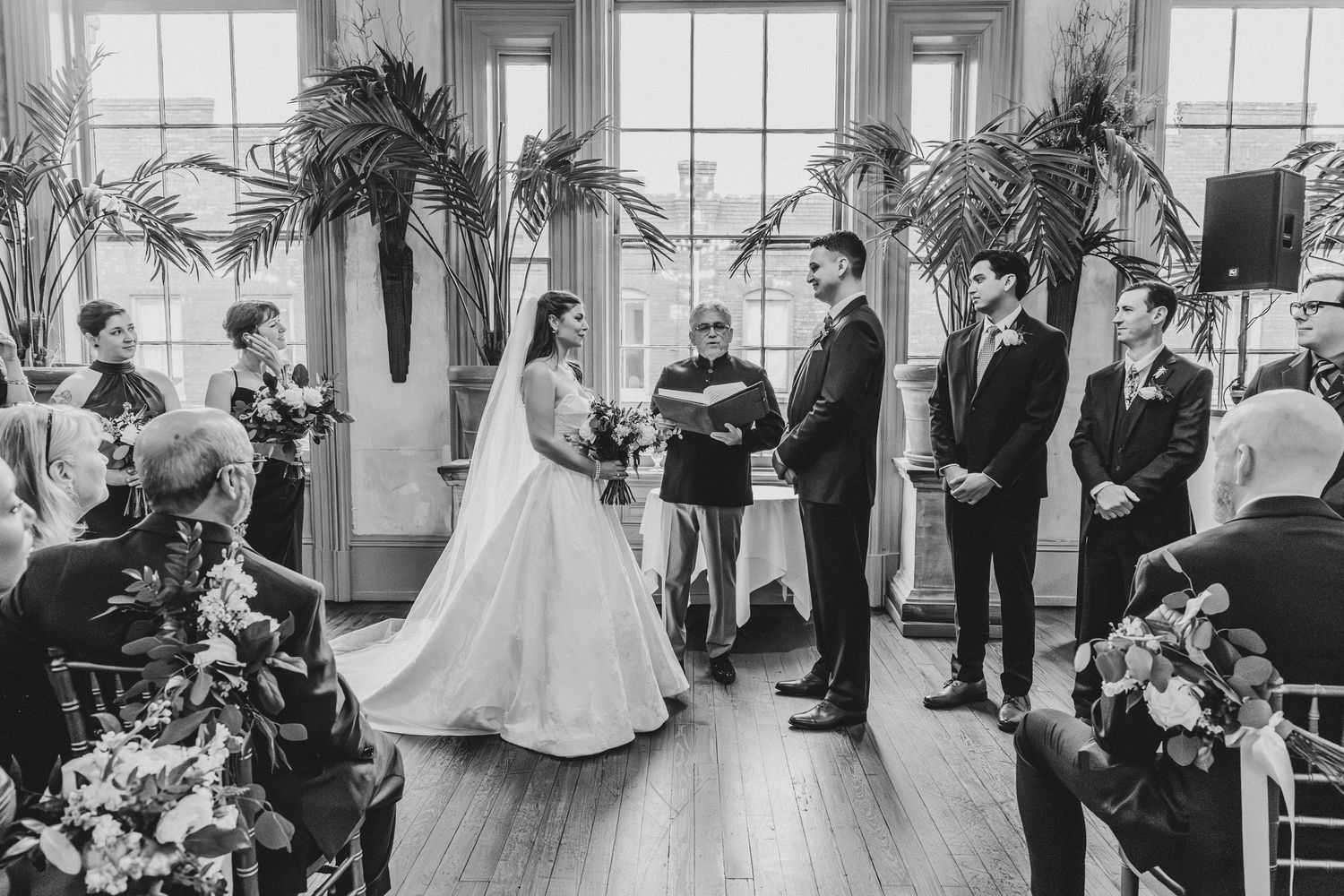 Black and white wedding ceremony photo with bride and groom at altar, officiant, groomsmen, and guests in elegant venue.