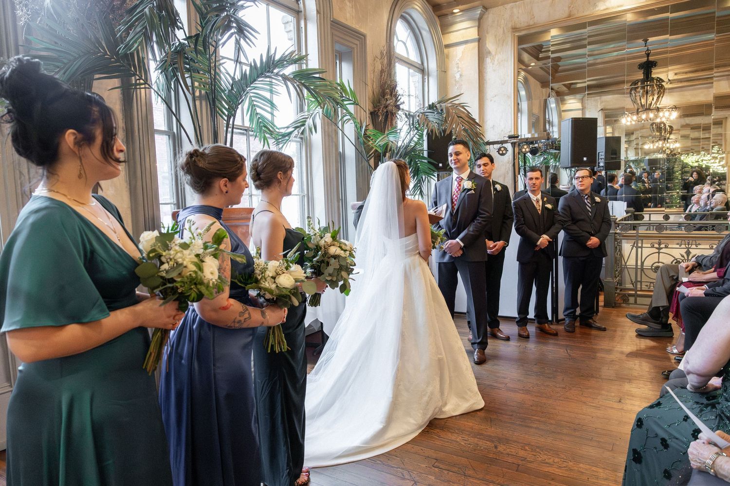 Elegant indoor wedding ceremony with bride in white gown, bridesmaids in blue and green, and groomsmen in suits.