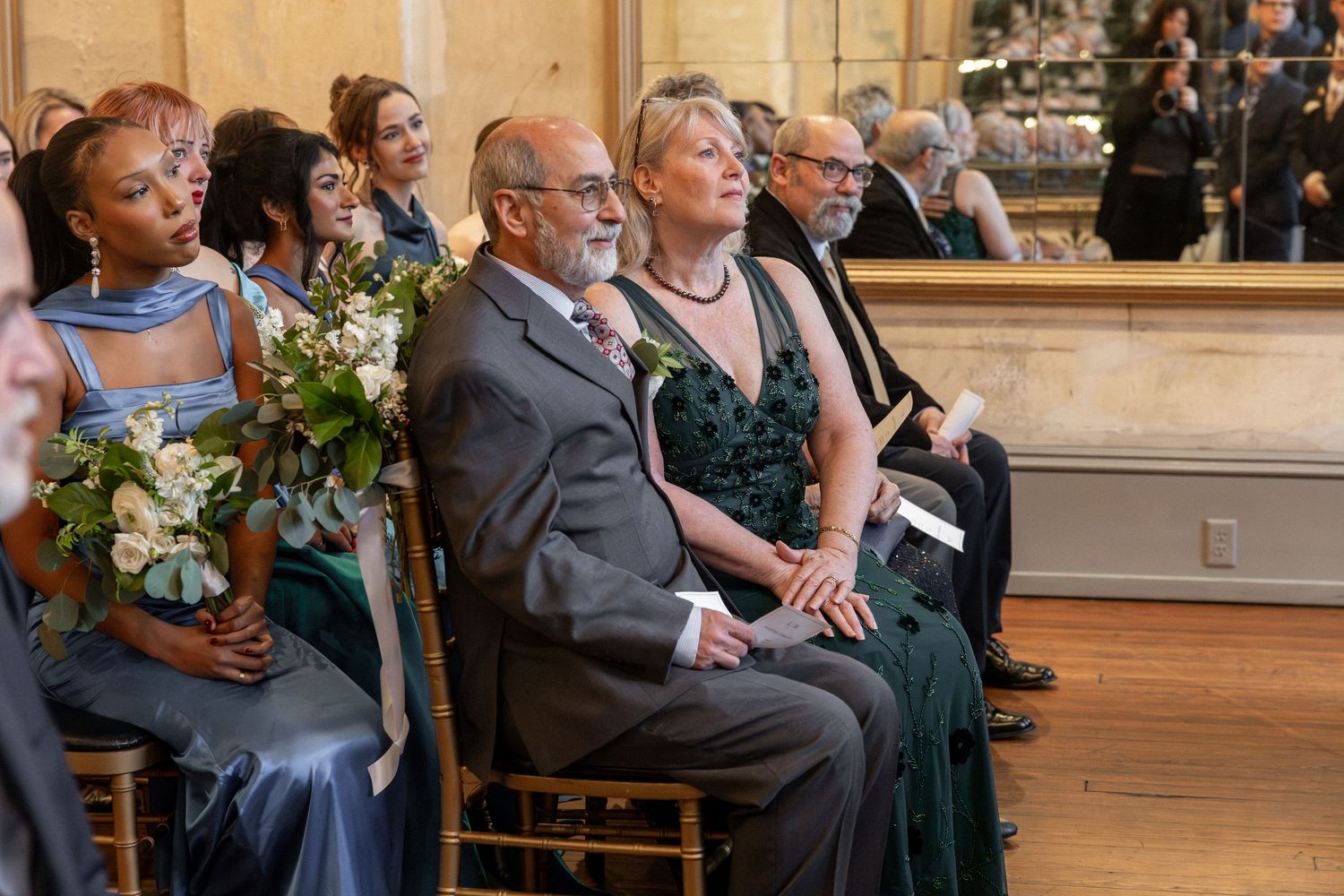 Guests seated at a wedding ceremony, wearing elegant green and blue attire, holding bouquets in a grand venue.