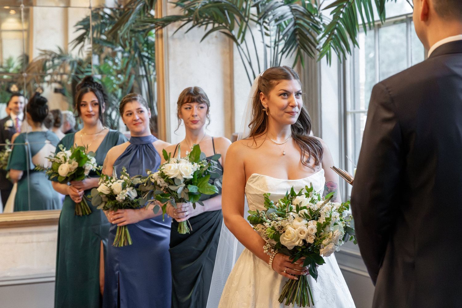Bride in white strapless gown holds bouquet with bridesmaids in blue and purple dresses at elegant wedding ceremony.