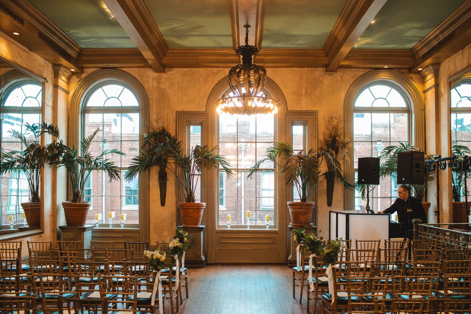 Elegant wedding ceremony venue with arched windows, chandelier, chiavari chairs, and tropical plants in a warmly lit ballroom.