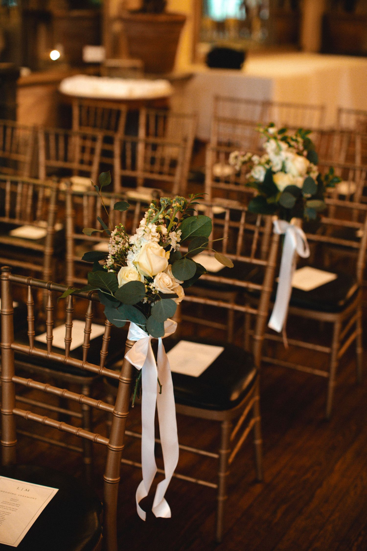 Gold chiavari chairs adorned with white rose bouquets and ribbon bows line a wedding ceremony aisle in an elegant venue.