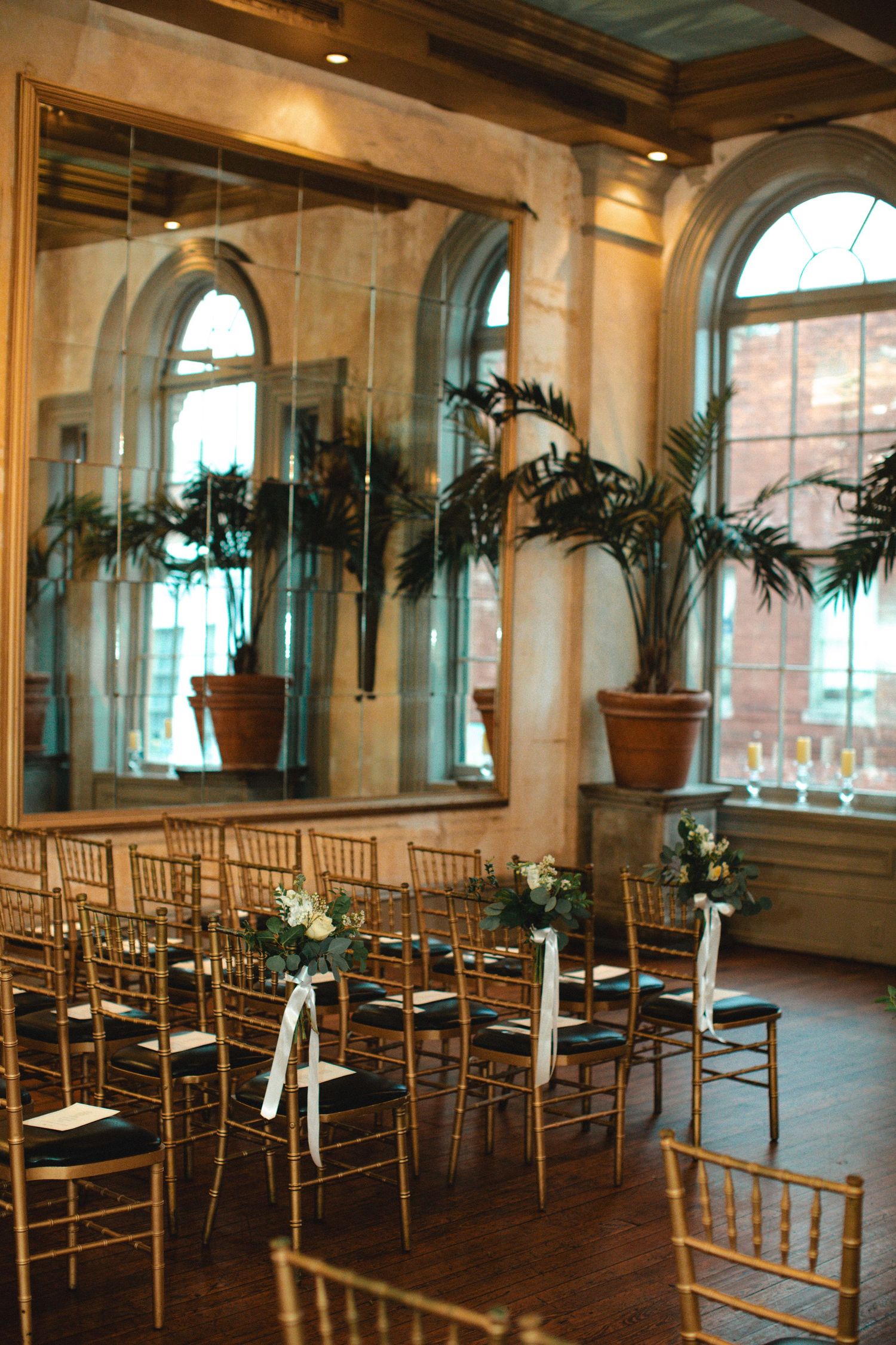 Elegant wedding ceremony setup with gold chiavari chairs, floral accents, large mirror, and tropical plants in a classic venue.