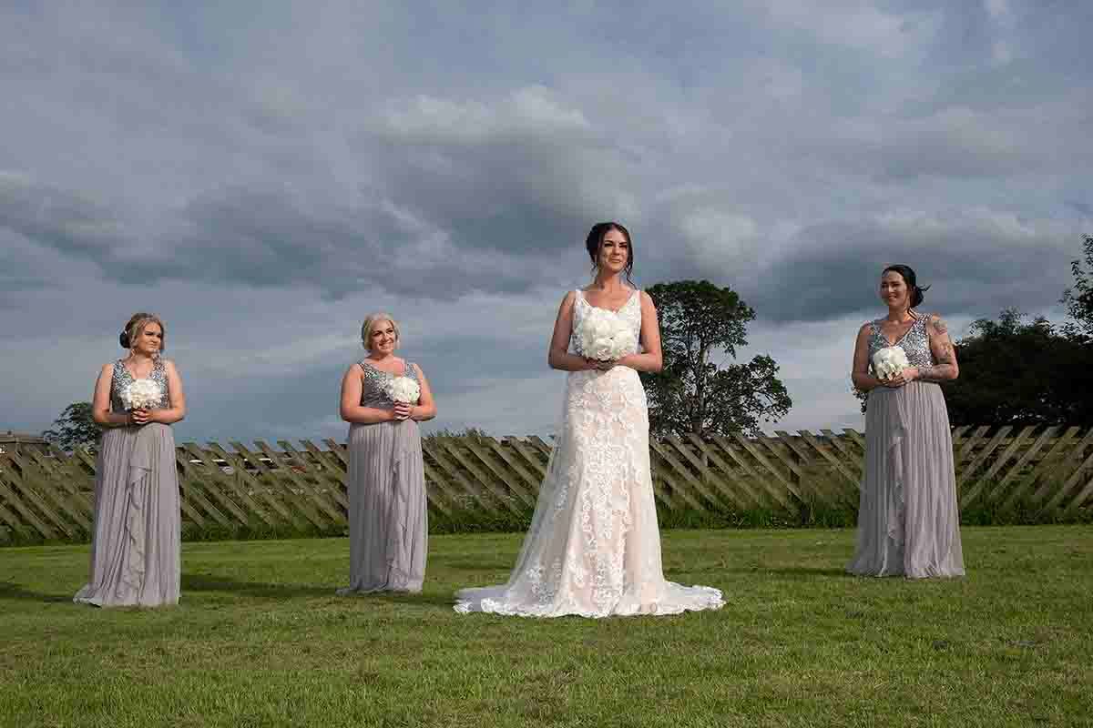 Bride in white lace gown stands with three bridesmaids in grey dresses holding bouquets on green lawn at Greenhill Hotel - Wigton- Cumbria