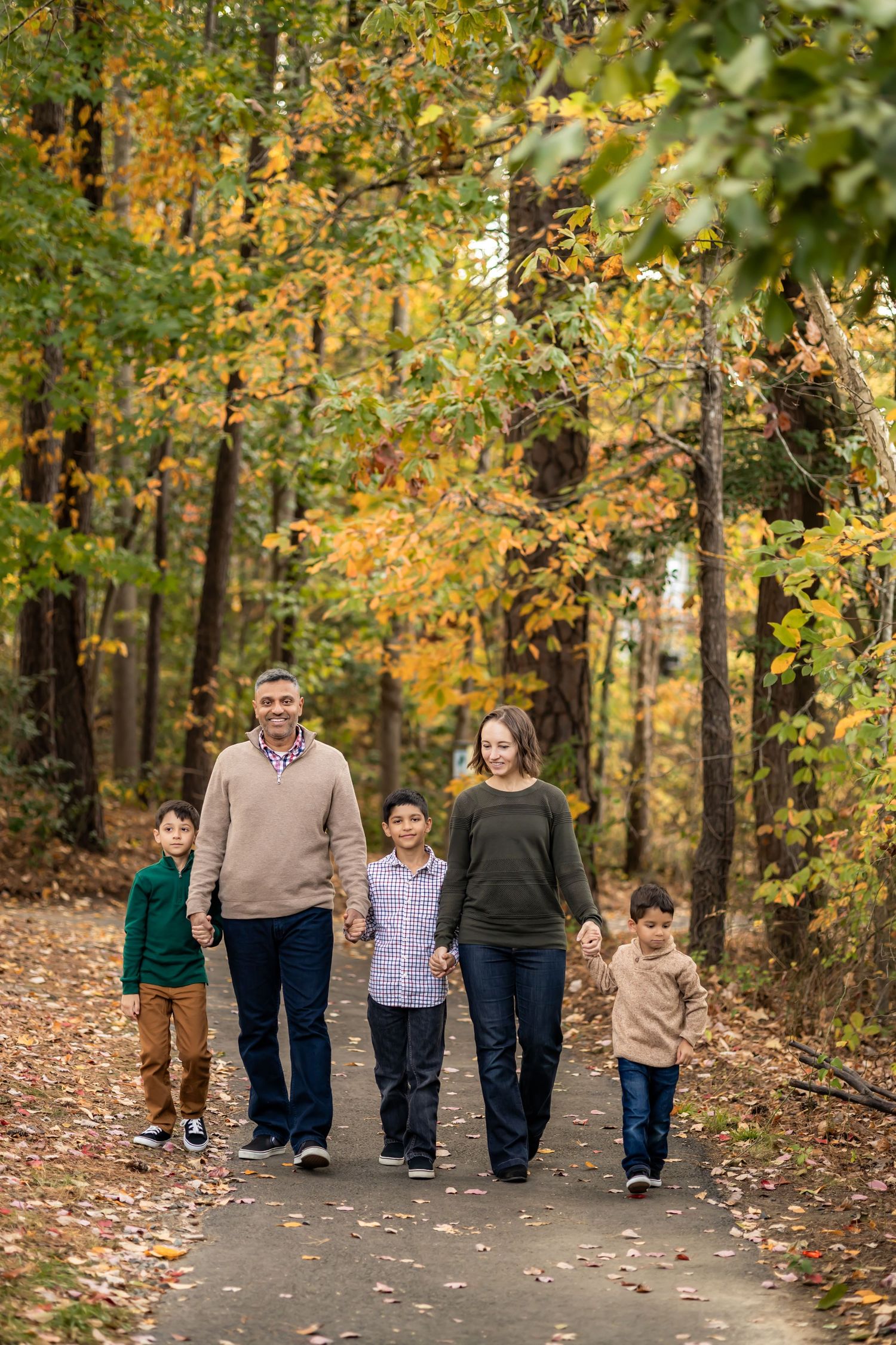 Family of five walking hand-in-hand on a tree-lined path surrounded by vibrant autumn foliage.