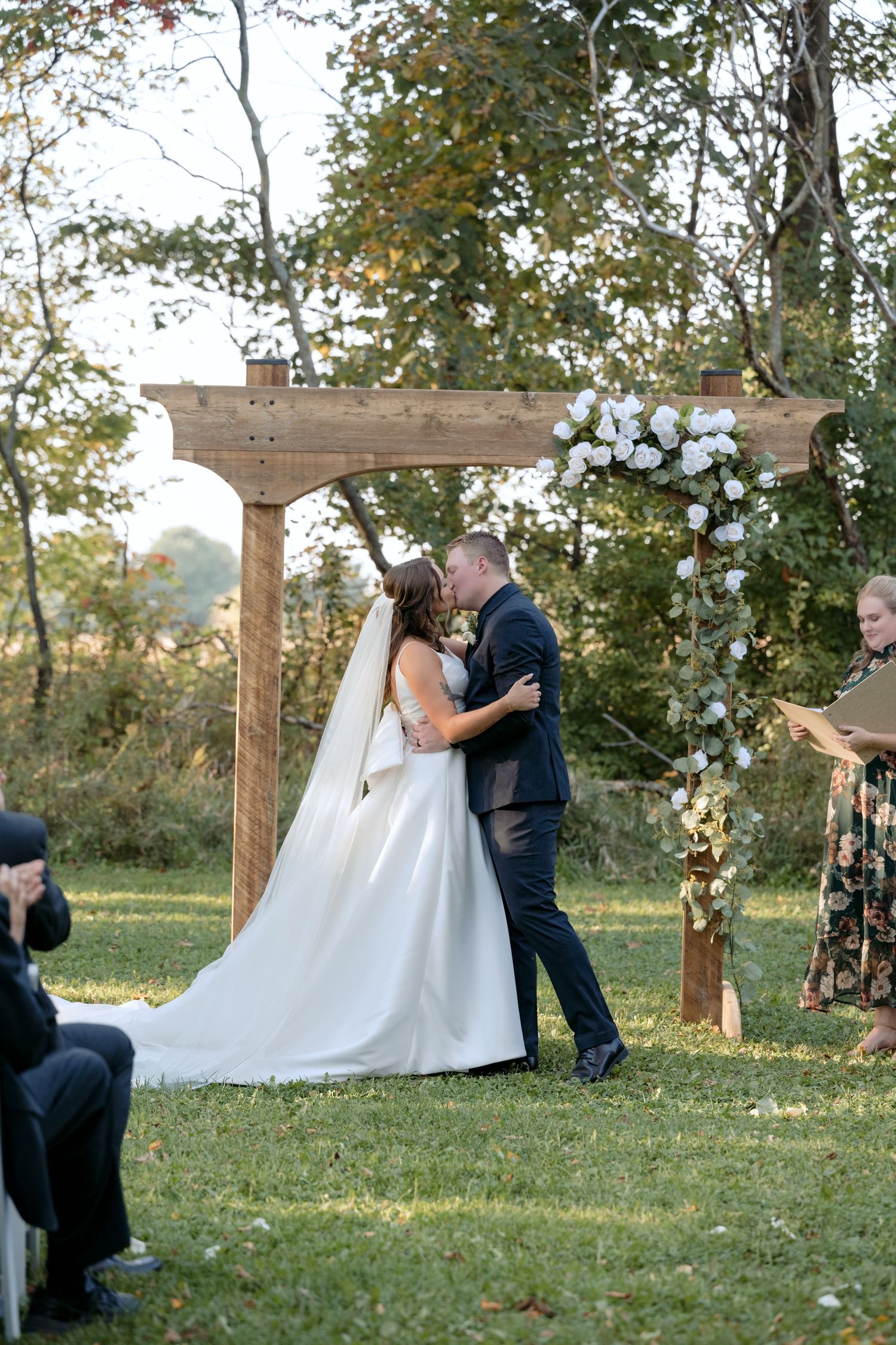 Bride and groom kiss during their wedding ceremony under a decorate arbor, surrounded by greenery. Photographed at Abella in Chisago City MN