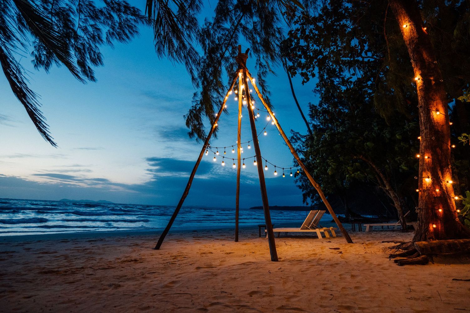 Tropical beachside restaurant with palm trees, warm string lights, and wooden tables at dusk.