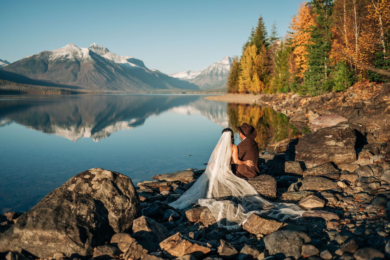 Bride and groom sit on rocky lakeshore with mountain reflections and autumn foliage at Glacier National Park.