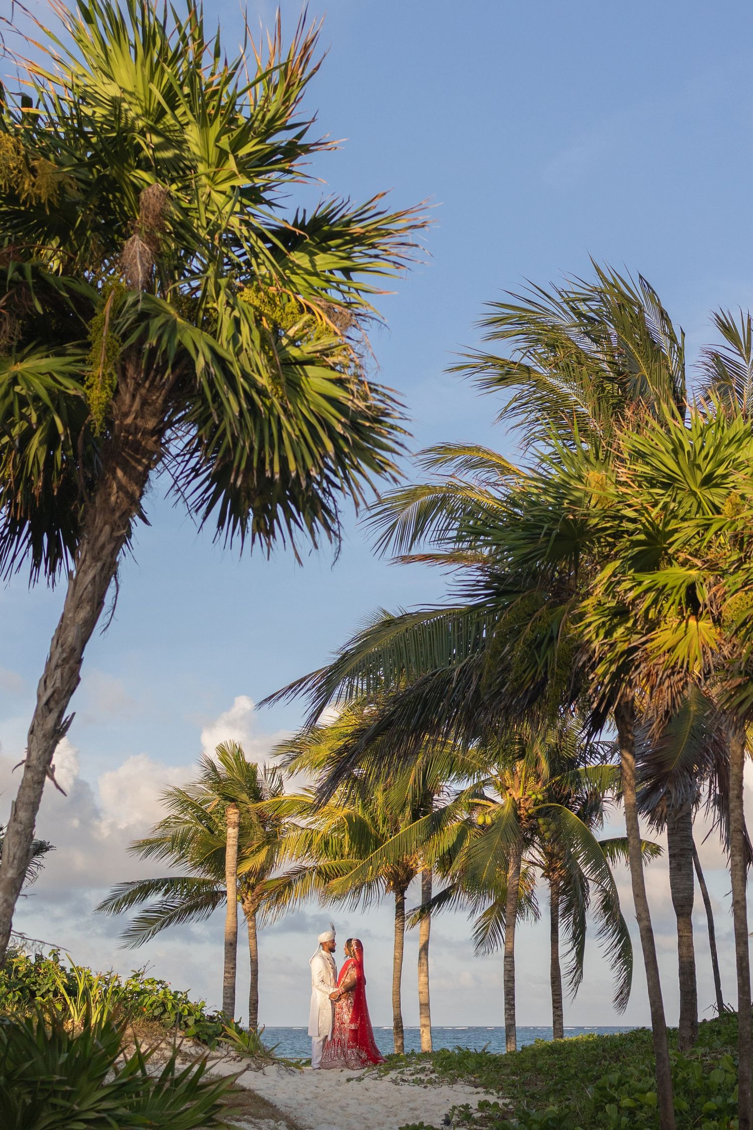 Couple in traditional attire standing among tropical palm trees on a sandy beach with blue sky backdrop.