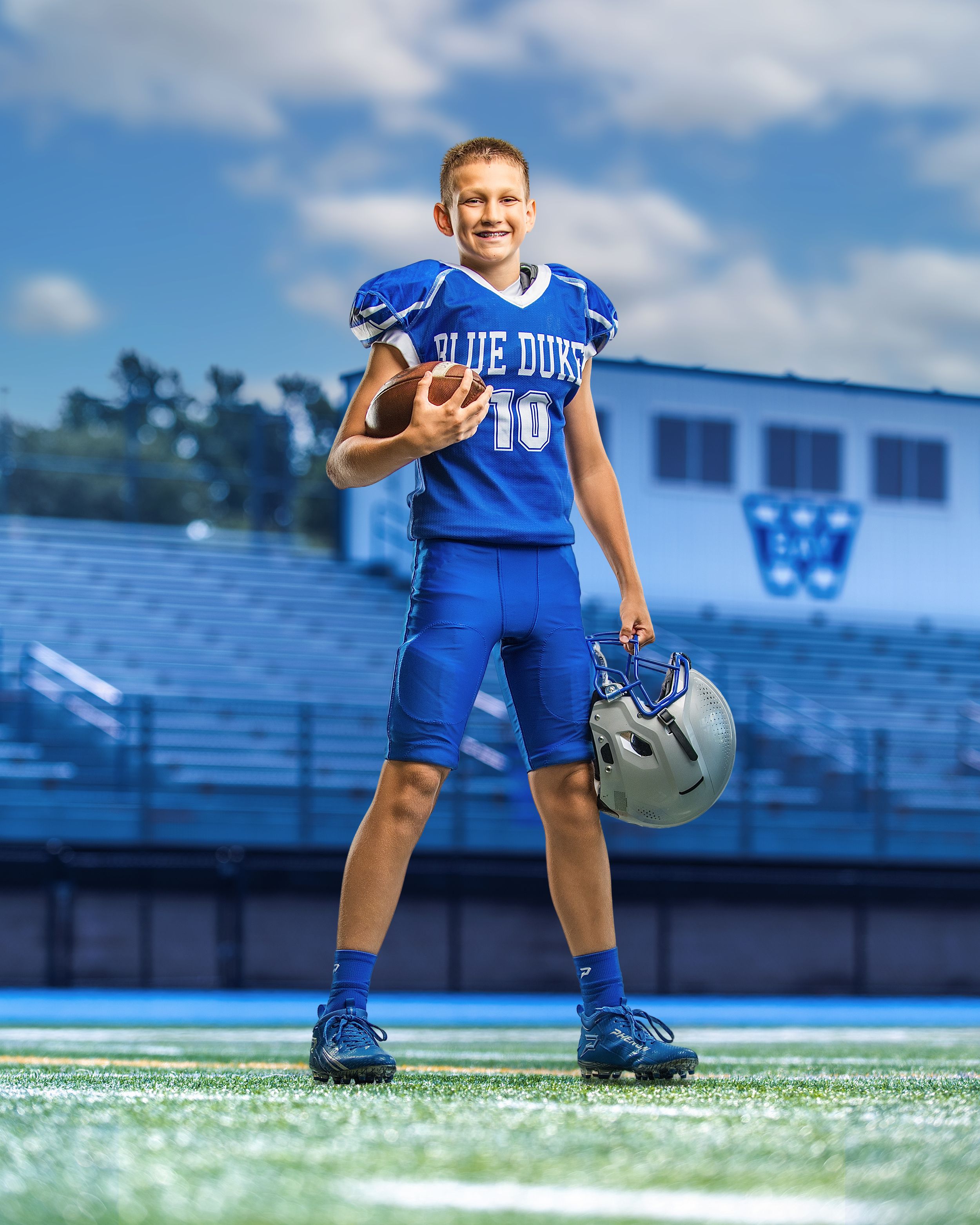 Young boy in Blue Dukes #10 football uniform holding helmet and ball on field.