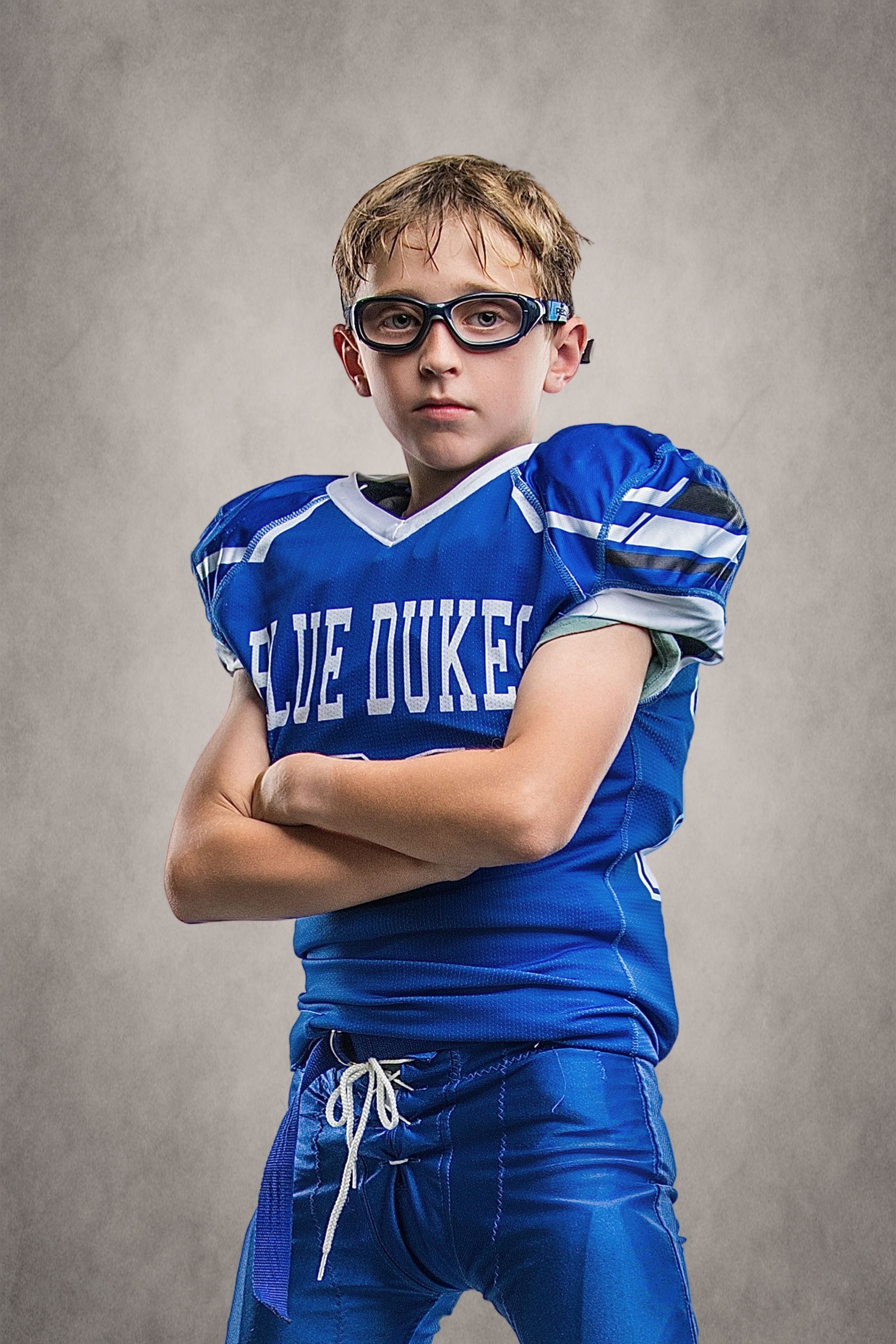 Young boy in blue Blue Dukes football jersey and glasses poses with arms crossed in a confident stance.