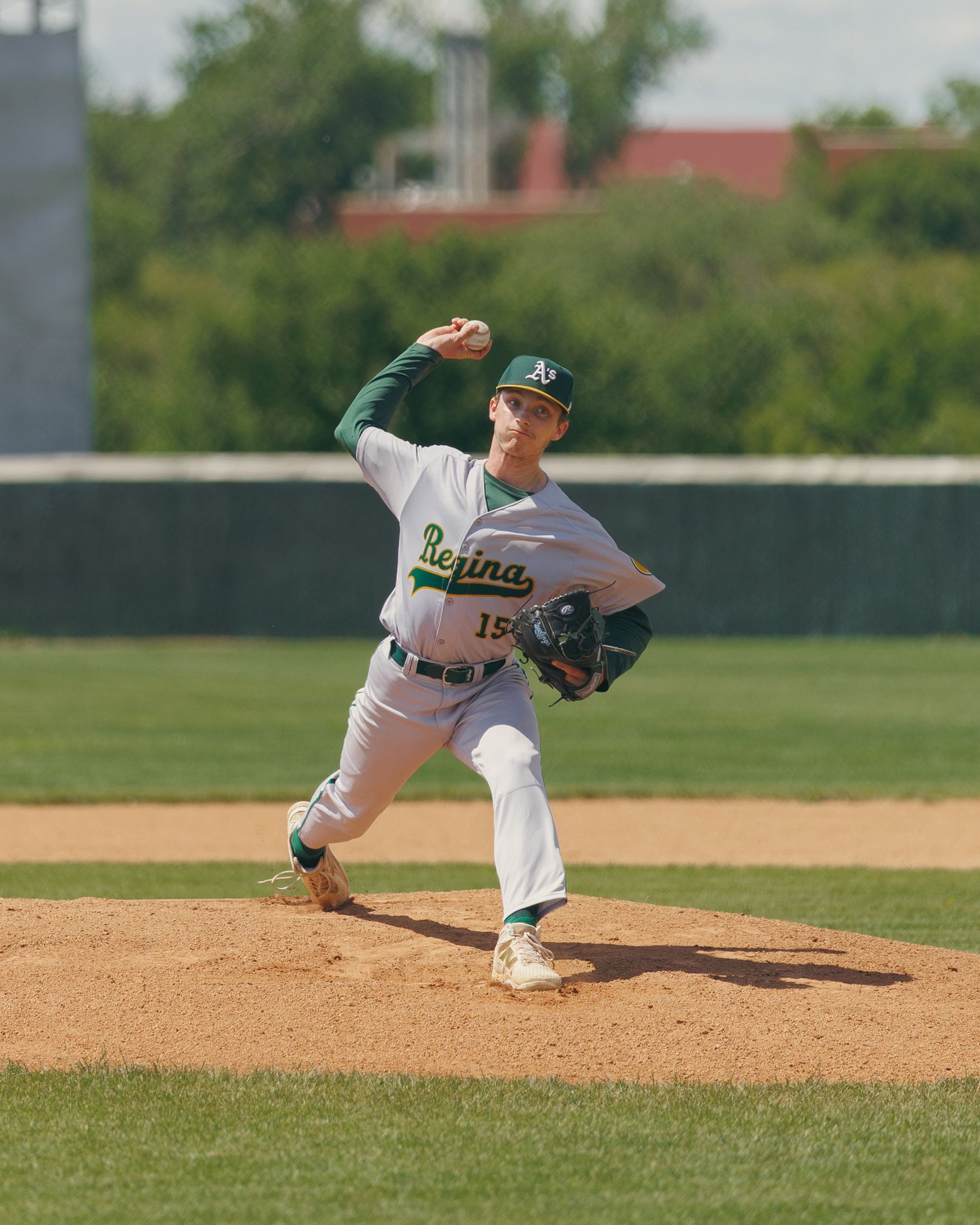 Regina baseball pitcher #15 winds up on mound in green and white uniform.