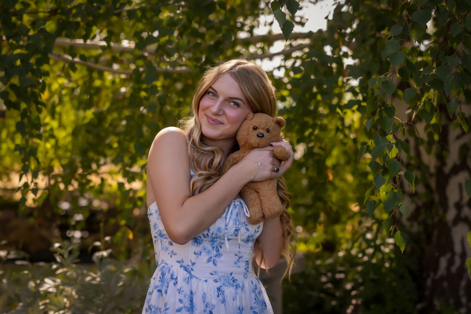senior girl in backyard wearing blue and white dress holding childhood teddy bear