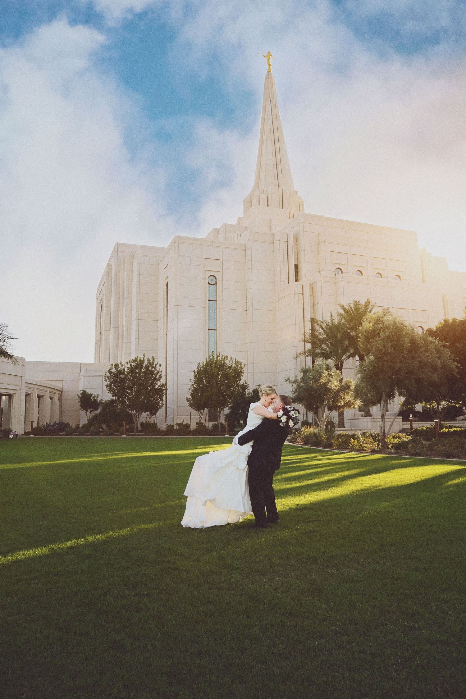 Bride in a white gown embraces her groom on a sunlit green lawn in front of a white temple with a tall spire.