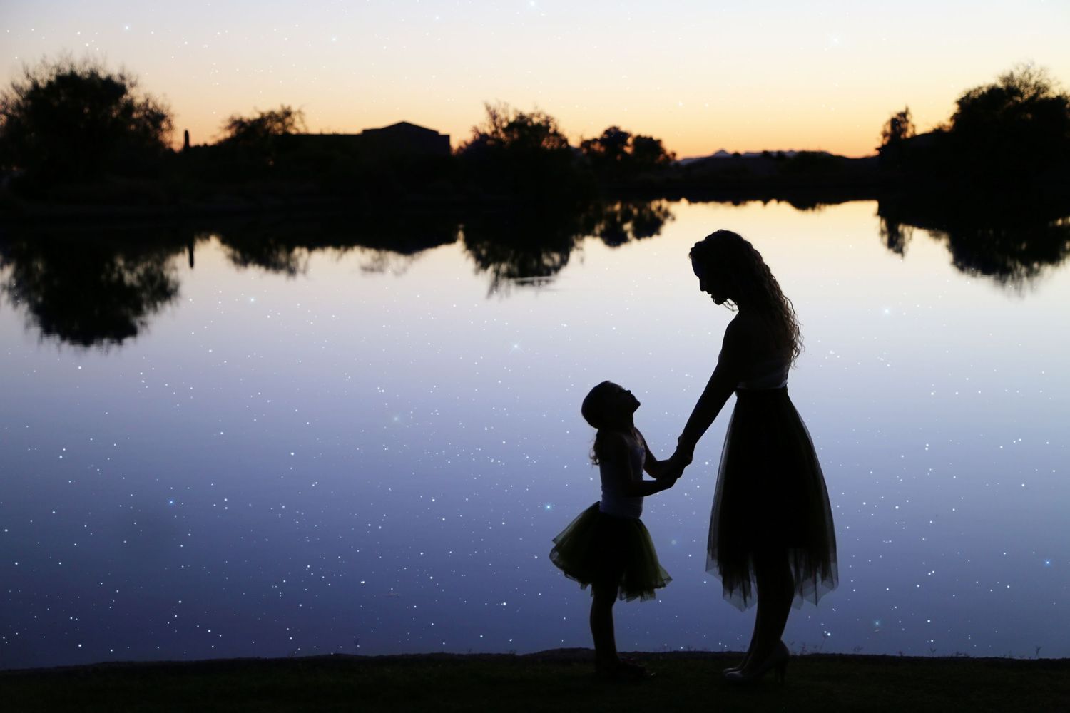 Silhouette of a mother and young daughter in a tutu skirt holding hands beside a calm lake at sunset.