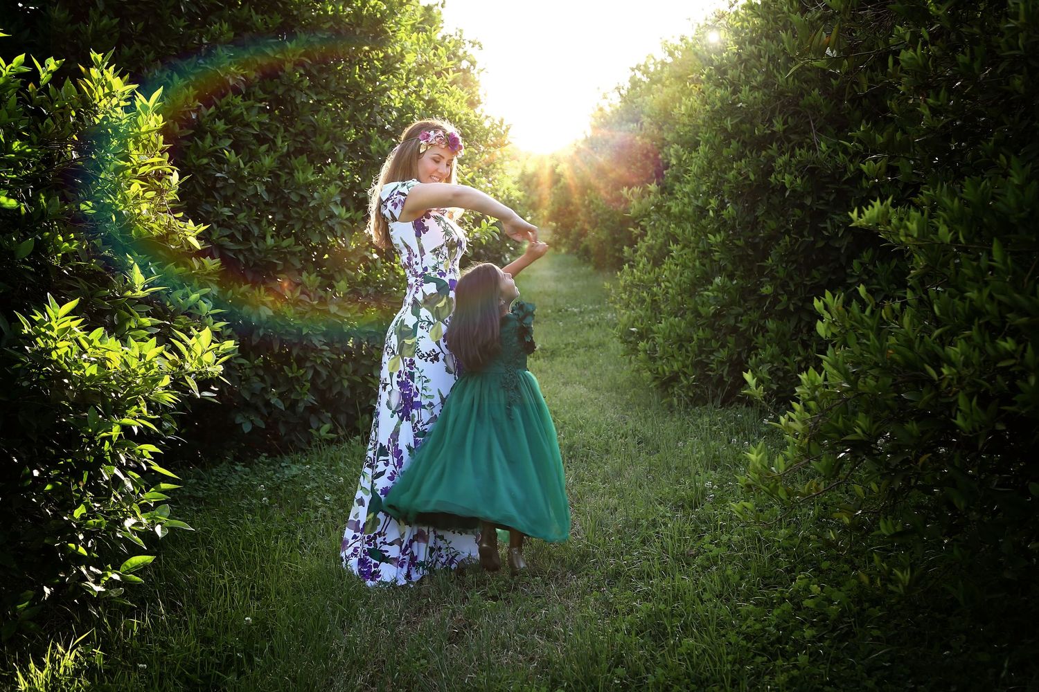 Mother in a floral maxi dress twirls her daughter in a green skirt along a grassy path lined with sunlit trees.