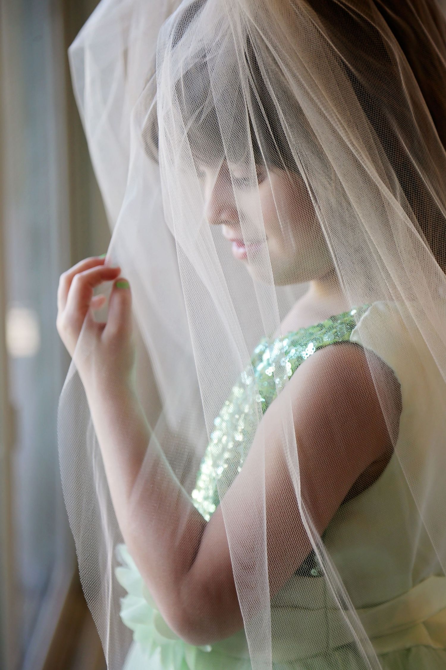 Young girl in a green sequined dress gazes down, a sheer tulle veil draped over her head, standing by a window.