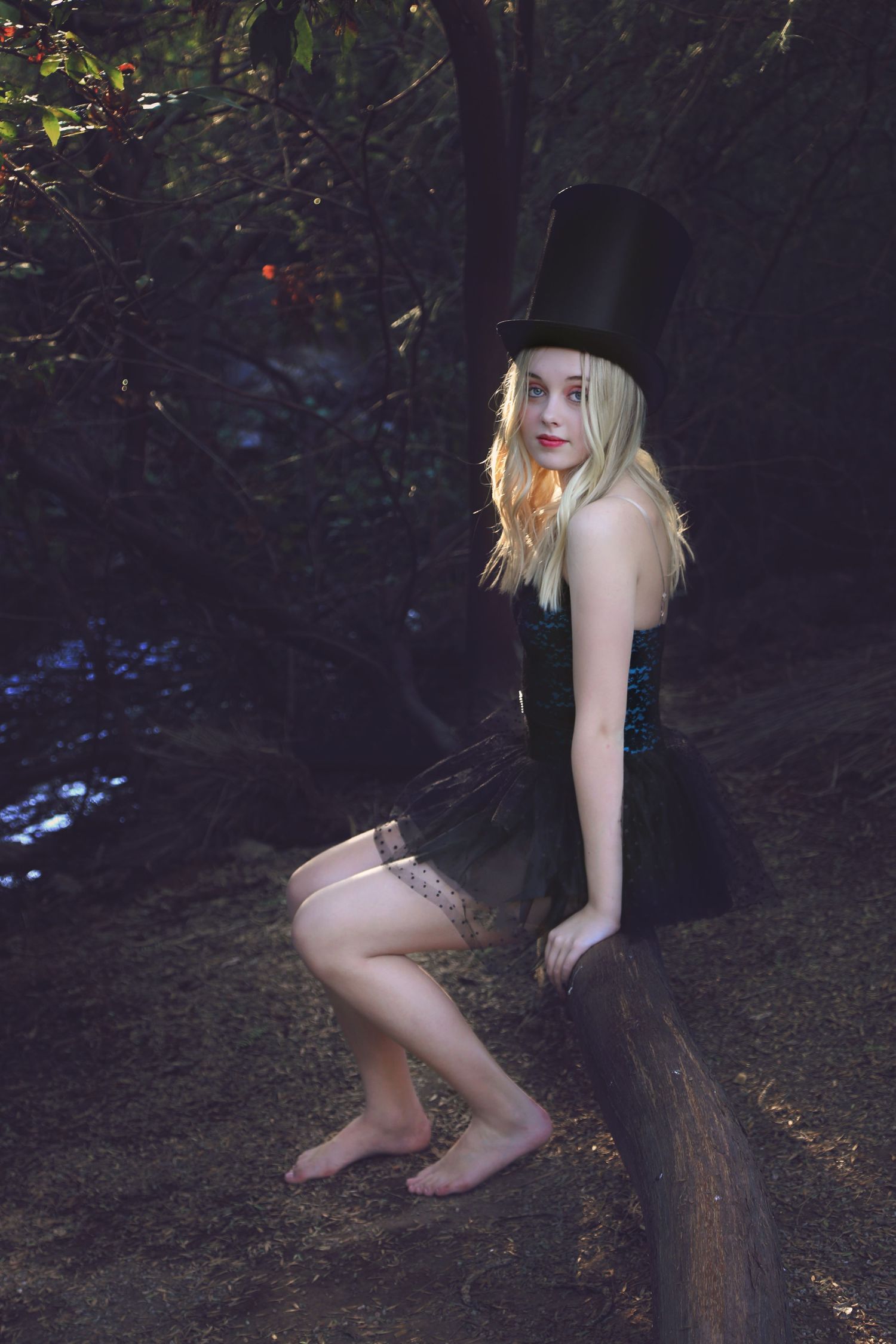Barefoot young woman in a black top hat and tulle skirt sits on a log in a dark, moody forest setting.