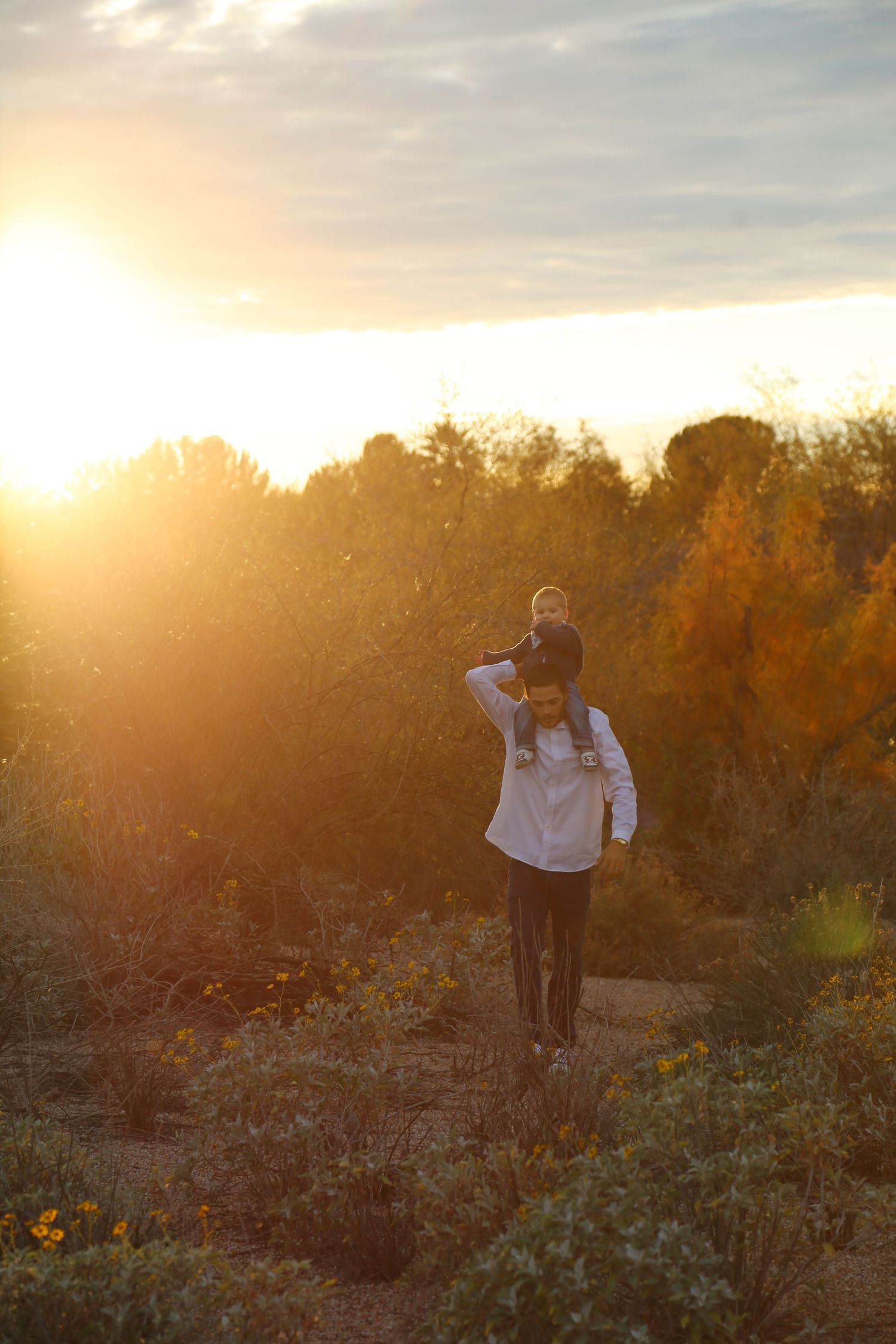 Parent carries a child on their shoulders along a dirt path through autumn shrubs at golden sunset.