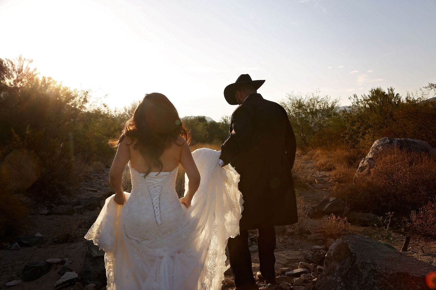 Bride in a white lace gown walks with a groom in a dark coat and cowboy hat through a rocky desert at sunset.