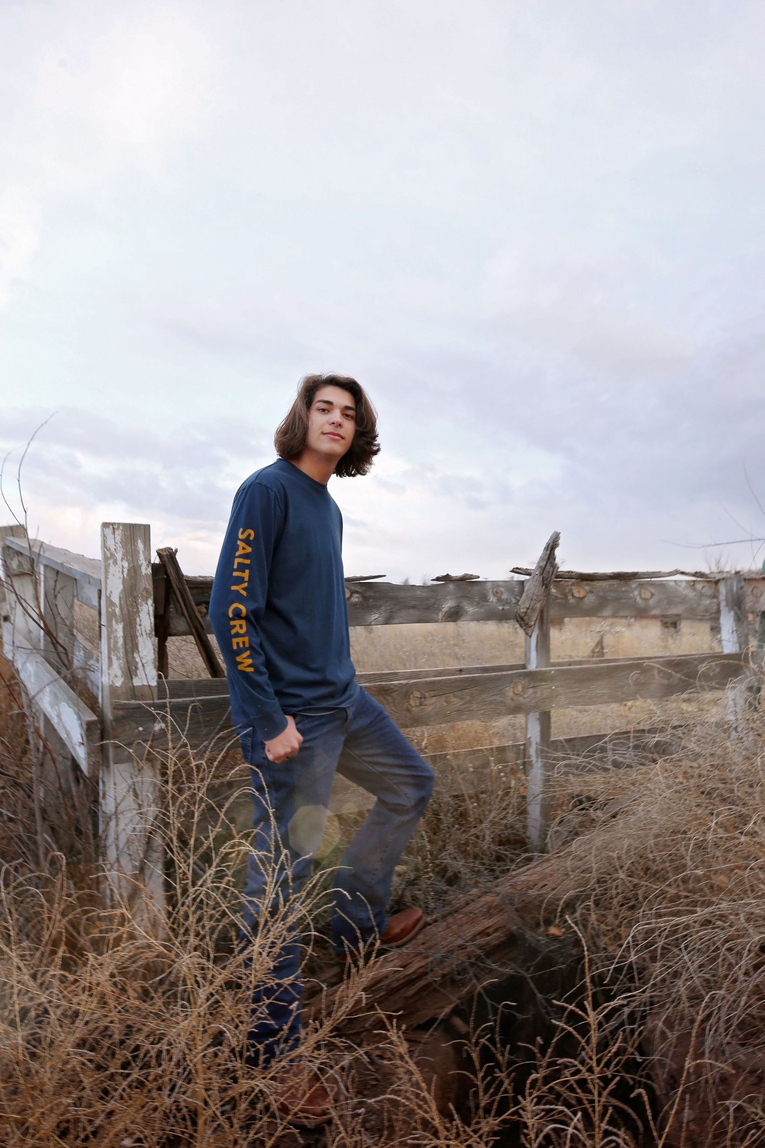 Teenage boy with wavy hair in a navy Salty Crew sweatshirt leaning on a weathered wooden fence under a cloudy sky.