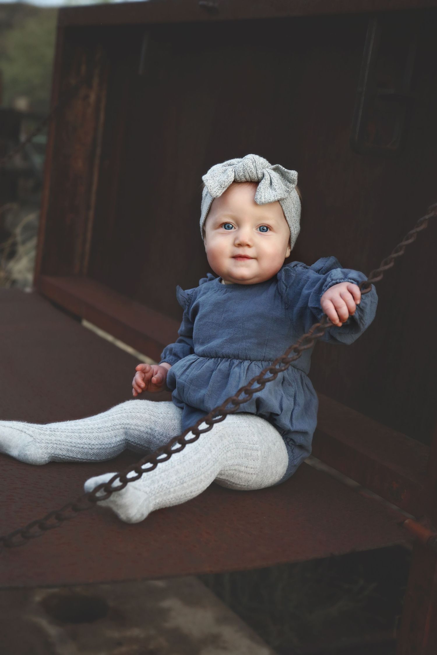 Smiling baby in a gray bow headband, blue ruffled dress and cream knit tights sitting on a rusty metal surface with a chain.