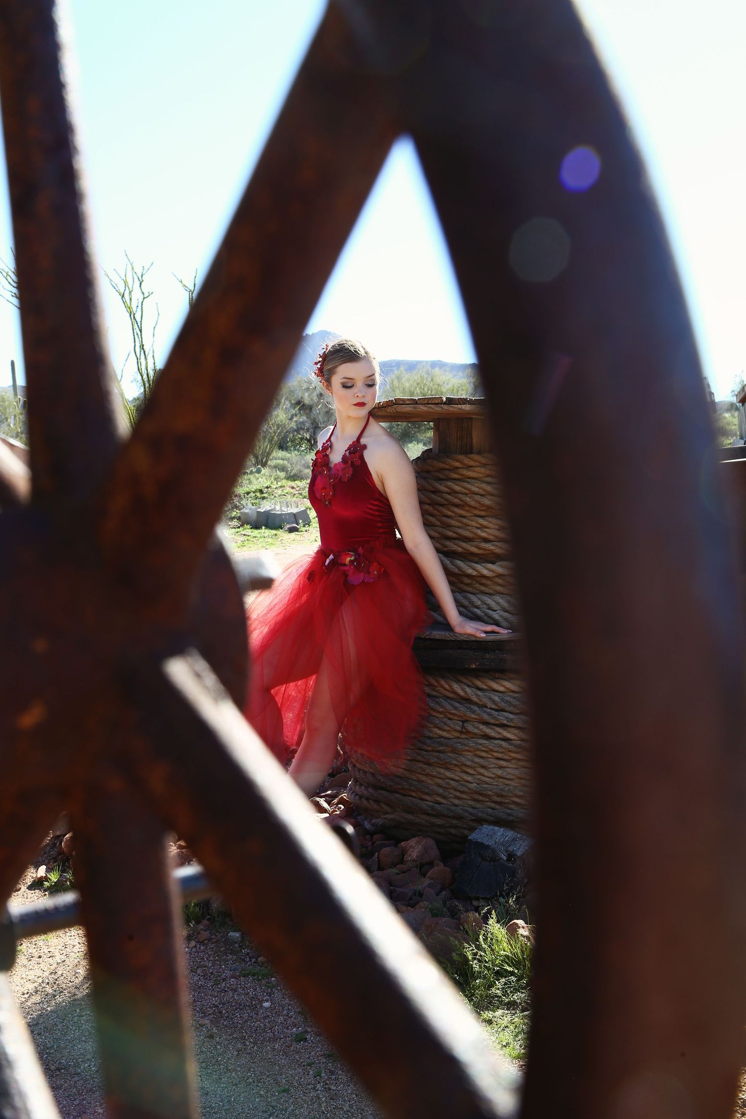Woman in a red tulle dress leans against a rope-wrapped barrel in the desert, framed by a rusty wagon wheel.
