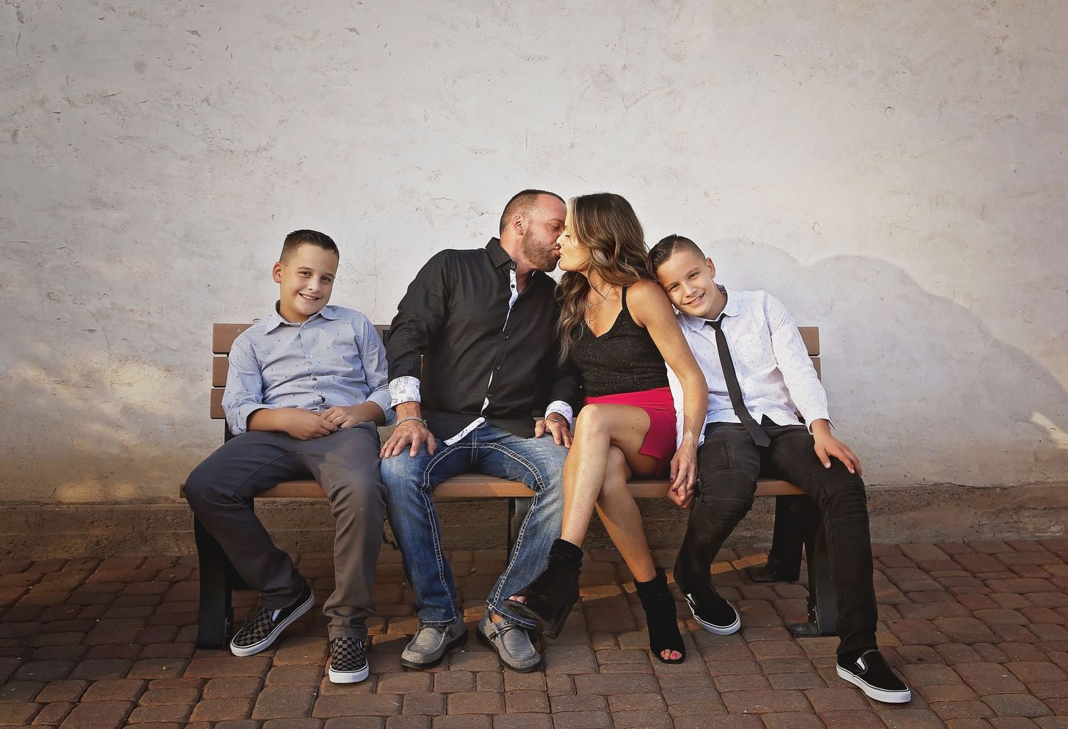 Family of four sits on a wooden bench against a rustic wall as two boys smile while their parents share a kiss.