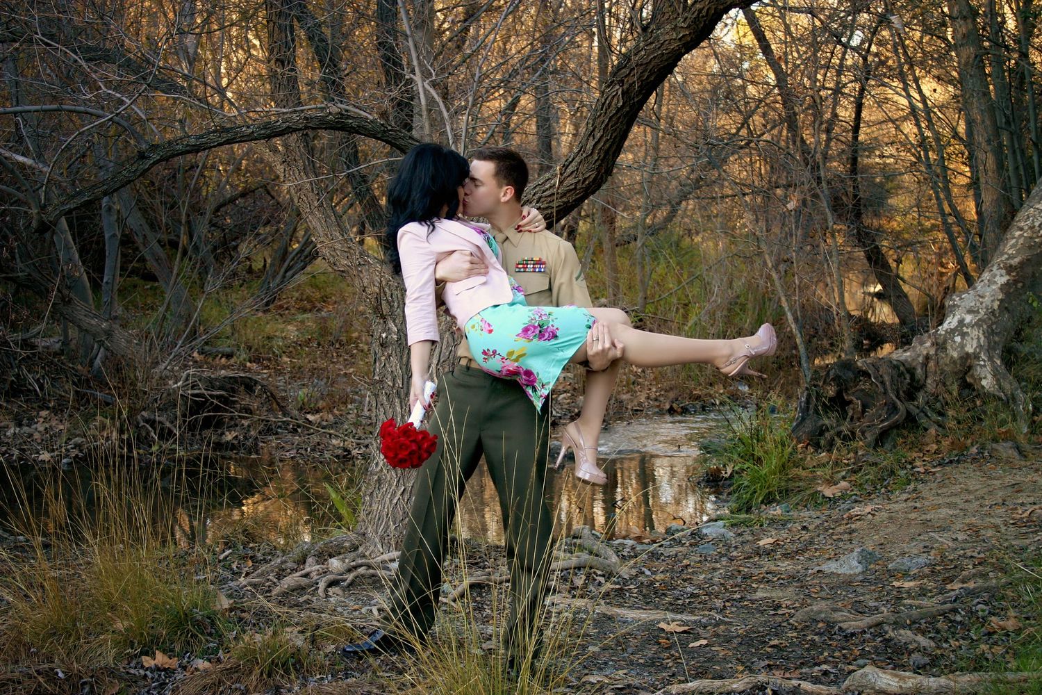 Man in a cowboy-style outfit carries a woman in a floral dress in his arms as they kiss in an autumn forest.